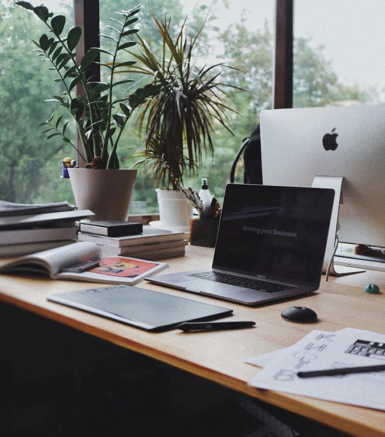 A tidy desk with a laptop, notebooks and plants by a window